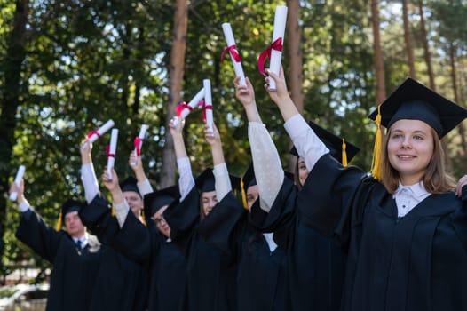 A group of graduates in robes raised their hands with diplomas outdoors. Elderly student