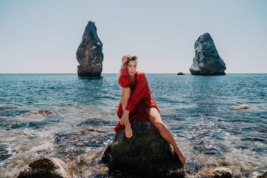 Woman travel sea. Young Happy woman in a long red dress posing on a beach near the sea on background of volcanic rocks, like in Iceland, sharing travel adventure journey