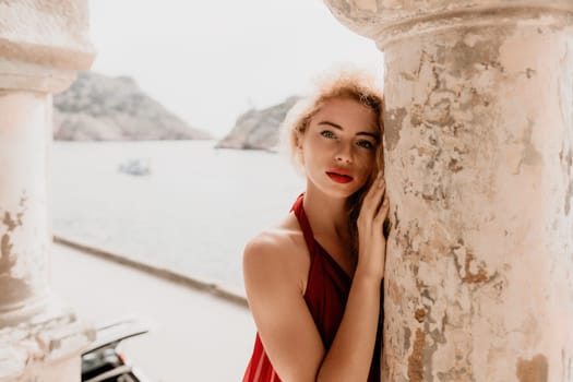 Close up shot of beautiful young caucasian woman with curly blond hair and freckles looking at camera and smiling. Cute woman portrait in a pink long dress posing on a volcanic rock high above the sea