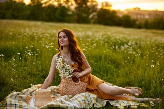 a happy woman in an orange dress sits on a plaid in a chamomile field at sunset and laughs. High quality photo