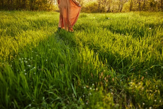 horizontal photo of a woman in an orange dress photographed without a face, standing in a green field during sunset, illuminated from the back. High quality photo