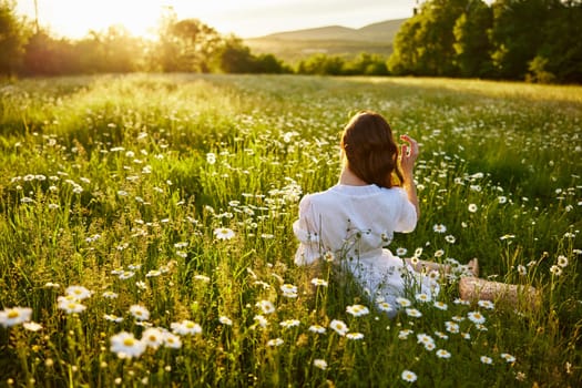 a red-haired woman in a light dress sits in a chamomile field at sunset and admires the passing day. High quality photo