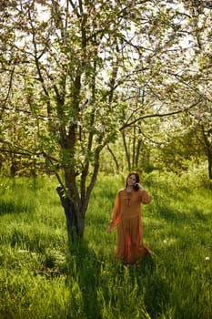 a beautiful, joyful woman stands in a long orange dress, in the countryside, near a tree blooming with white flowers, during sunset, illuminated from behind and looks into the camera. High quality photo