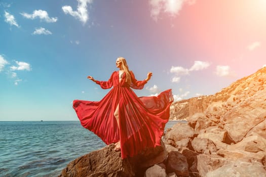 Red dress sea woman. A blonde with flowing hair in a long flowing red dress stands on a rock near the sea. Travel concept, photo session at sea.