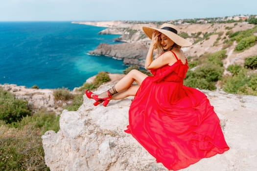A woman in a red silk dress sits by the ocean with mountains in the background, her dress swaying in the wind
