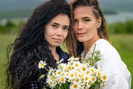 Two women enjoy nature in a field of daisies. Girlfriends hugging hold a bouquet of daisies and look at the camera