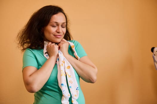 Beautiful Latin American pregnant woman hugging a cute baby jumpsuit, standing with her eyes closed over yellow background, expressing positive emotion, enjoying happy carefree pregnancy and maternity