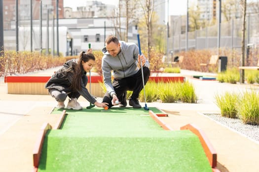 Sharing with golf experience. Cheerful young man teaching his daughter to play mini golf at the day time. Concept of friendly family.