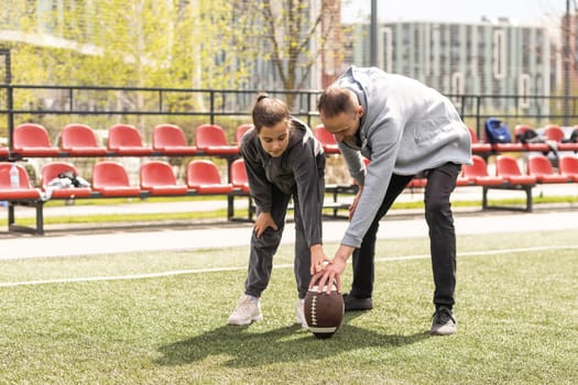 Handsome dad with his little cute daughter are having fun and playing American football on green grassy lawn.