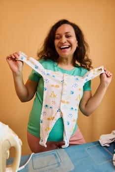 Overjoyed expecting adult woman with bodysuit for newborn baby, smiling cheerfully looking at camera, expressing positive emotions over orange isolated background. Happy moments of carefree pregnancy.