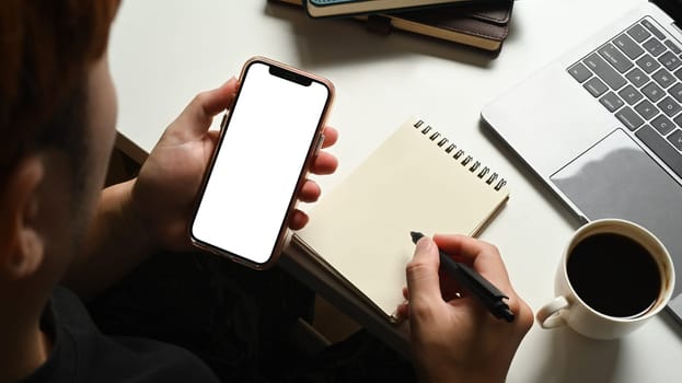 Closeup view of man holding mobile phone and making notes on notepad. Blank screen for advertising text message.