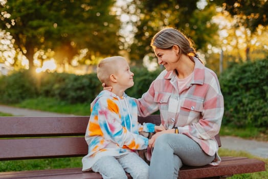 mother and son sit on a park bench in the rays of the setting sun. the concept of a family. Mother's Day. beautiful girl (mother) with a boy (son) in the park in the park are sitting on a bench at sunset.