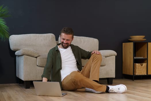 Man sitting on floor near sofa in house and working on laptop. He is freelancer or remote worker, using technology and internet to do job. High quality photo