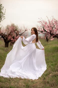 Woman peach blossom. Happy curly woman in white dress walking in the garden of blossoming peach trees in spring.