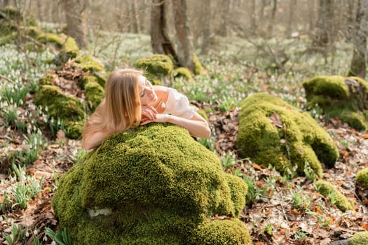 Snowdrops galanthus blond. A girl in a white dress lay down on a stone in the moss in a meadow with snowdrops in a spring forest.