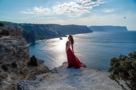 A girl with flowing hair in a long red dress stands on a rock above the sea. The stone can be seen in the sea. Sunny path to the sea from the sun