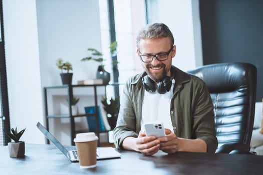 European man is taking break in office and using phone to send text message, illustrating concept of easy communication. Casual and relaxed work environment. High quality photo