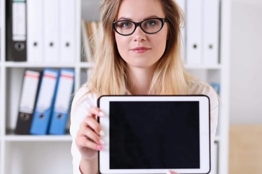 Beautiful businesswoman wearing glasses portrait at the office holding a tablet in hand sitting at the table smiling and looking at the camera teacher expresses success checking the test papers.