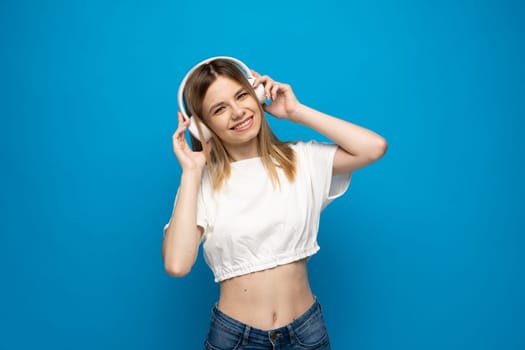 Young blonde woman in the white t-shirt with headphones listening music .Music teenager girl dancing against isolated blue background