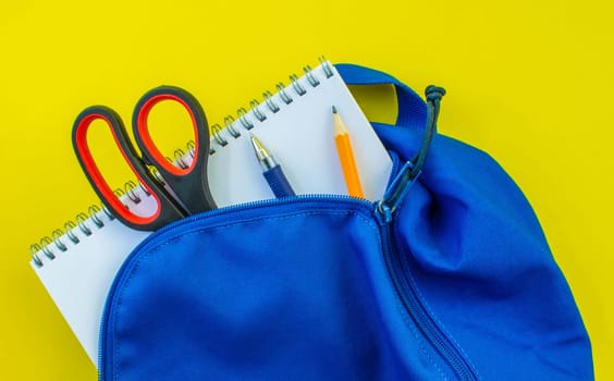 School backpack, pencil, pen, scissors and notepad on a yellow background. School blue backpack with notepad, pencil, scissors and pen inside on a yellow background.