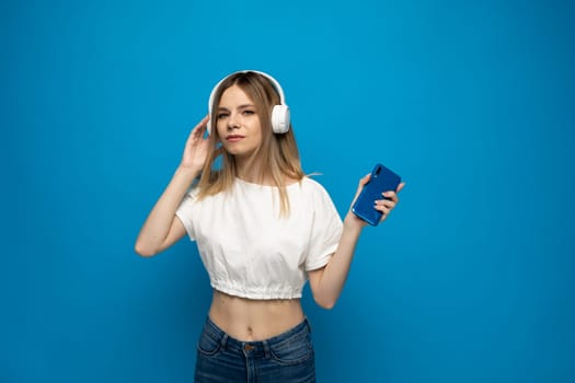 Young blonde woman in white t-shirt with headphones listening music. Music teenager girl dancing against isolated blue background
