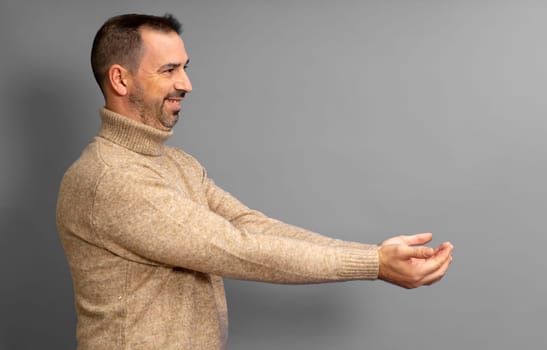 Side view portrait of friendly handsome bearded man offering something in his hands with outstretched arms. Indoor studio shot isolated on gray background.