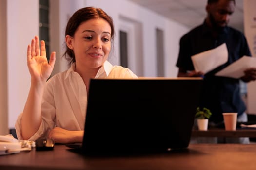 Smiling employee greeting colleague on videocall, discussing startup project, waving hello. Corporate worker answering teleconference call in office with sunset light, remote conversation