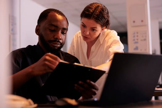 Colleagues discussing report statistics, comparing data on laptop in office. Two corporate workers checking marketing research results in coworking space with evening sunset light