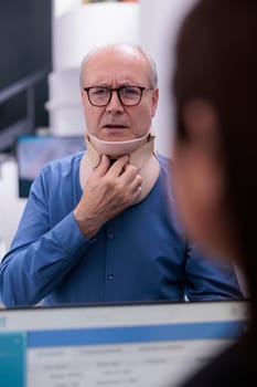 Elderly patient wearing cervical neck collar in hospital waiting room while asking receptionist about emergency care. Old man with medical brace waiting to meet with traumatologist.