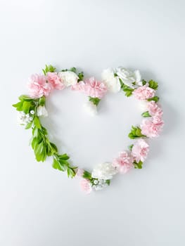 Frame in the form of a heart from pink and white carnations, green leaves, gypsophila on a white background. Flat lay, top view. Spring background. Suitable for wedding, cards and invitations