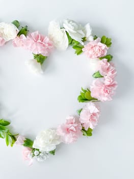 Frame in the form of a heart from pink and white carnations, green leaves, gypsophila on a white background. Flat lay, top view. Spring background. Suitable for wedding, cards and invitations