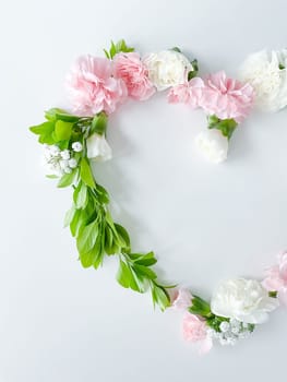 Frame in the form of a heart from pink and white carnations, green leaves, gypsophila on a white background. Flat lay, top view. Spring background. Suitable for wedding, cards and invitations