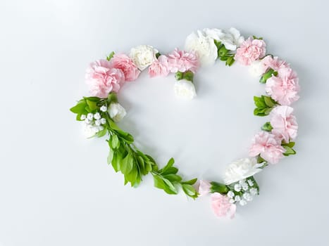 Frame in the form of a heart from pink and white carnations, green leaves, gypsophila on a white background. Flat lay, top view. Spring background. Suitable for wedding, cards and invitations