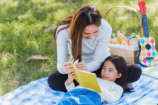 Happy Asian young family mother and child little girl having fun and enjoying outdoor laying on picnic blanket reading book at summer garden spring park, Family relaxation concept