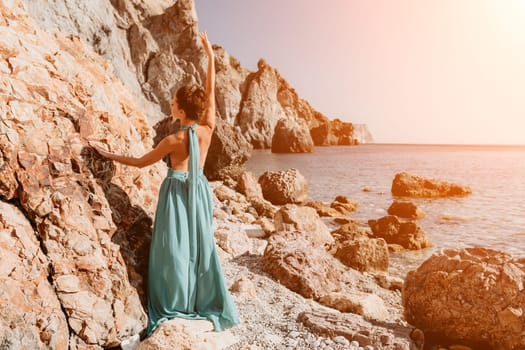 Side view a Young beautiful sensual woman in a red long dress posing on a rock high above the sea during sunrise. Girl on the nature on blue sky background. Fashion photo.