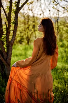 a lovely woman stands in nature in a bright, long orange dress, illuminated from the back by the sunset rays of the sun and holds the skirt of the dress with her hand, looking to the side enjoying the day. High quality photo