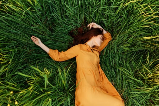 an elegant woman lies in the tall green grass in a long summer orange dress and with her arms outstretched enjoys nature and sunny weather with her eyes closed. High quality photo