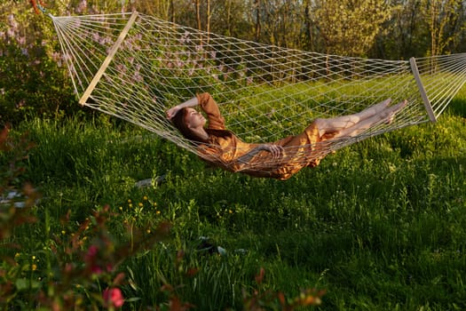 a happy woman in a long orange dress is relaxing in nature lying in a mesh hammock enjoying summer and vacation. High quality photo