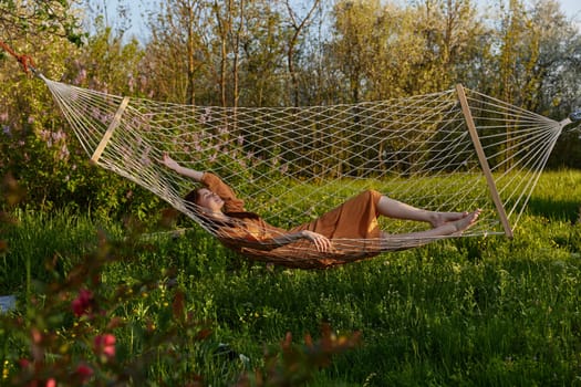 a happy woman in a long orange dress is relaxing in nature lying in a mesh hammock enjoying summer and vacation. High quality photo