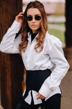 A stylish girl in a white shirt and sunglasses standing on the street during the day.