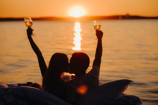 Silhouettes of a happy couple raising glasses on a summer evening near the sea at sunset.