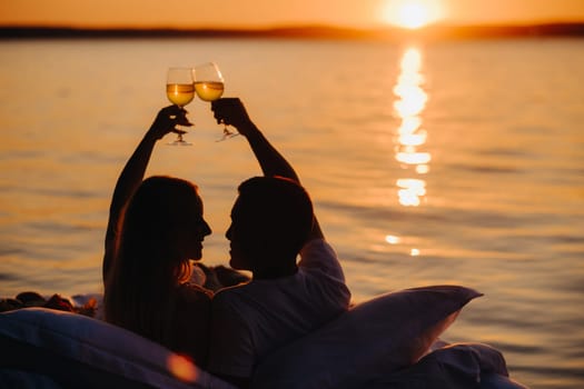 Silhouettes of a happy couple raising glasses on a summer evening near the sea at sunset.
