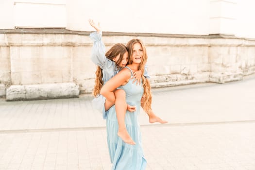 Mother of the daughter walks playing. Mother holds the girl on her back, holding her legs, and her daughter hugs her by the shoulders. Dressed in blue dresses