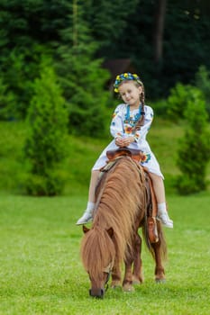 A little girl in the Ukrainian national costume rides a pony on the lawn