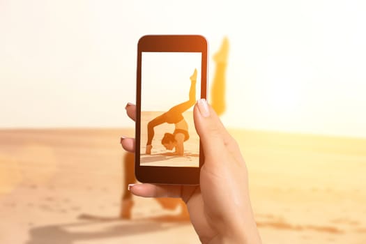 Image of female hands holding mobile phone with photo camera mode on the screen. A one woman doing acrobatic stunt on sand.