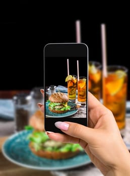 Photographing food concept - woman takes picture of sandwich with chicken burger, cheese and lettuce. Burger is on the table with paper and salt and pepper shakers. As well as two cocktails log Island Iced