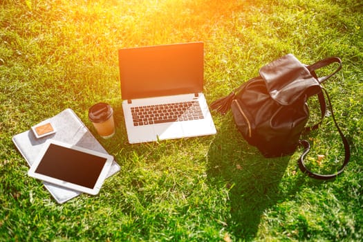 Laptop computer on green grass with coffee cup, bag and tablet in outdoor park. Copy space. Still life. Sun flare