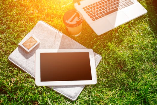 Laptop computer on green grass with coffee cup, bag and tablet in outdoor park. Copy space. Still life. Sun flare