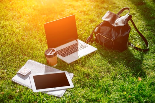 Laptop computer on green grass with coffee cup, bag and tablet in outdoor park. Copy space. Still life. Sun flare