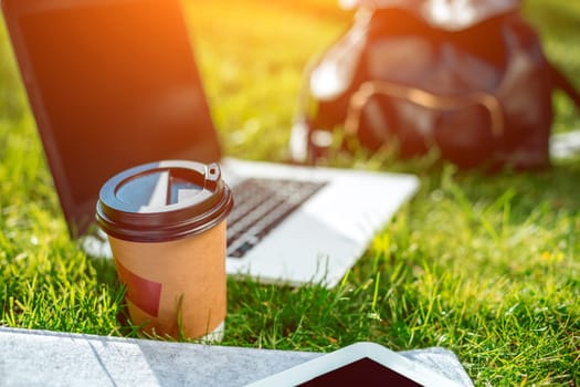 Laptop computer on green grass with coffee cup, bag and tablet in outdoor park. Copy space. Still life. Sun flare